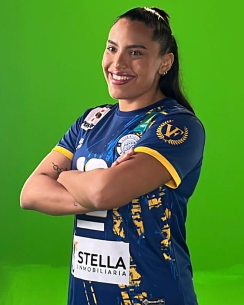 A confident female volleyball player, smiling broadly, stands with her arms crossed in front of a green screen. She has dark hair pulled back in a ponytail and is wearing a dark blue and yellow short-sleeved jersey with a sponsor logo "STELLA INMOBILIARIA" on the front, and dark blue athletic shorts. The jersey also features various patterns and emblems.